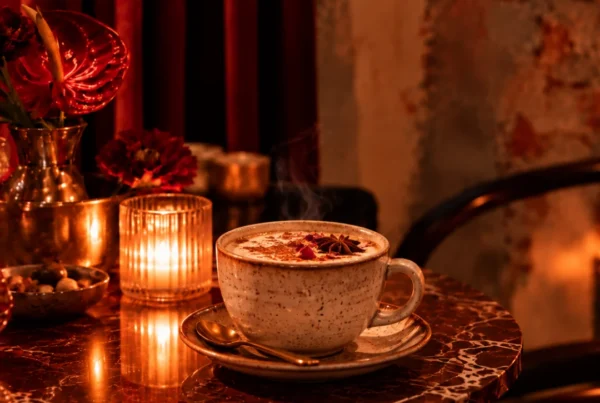 Speckled mug of chai on saucer beside a lit candle and brass vase with red flowers.