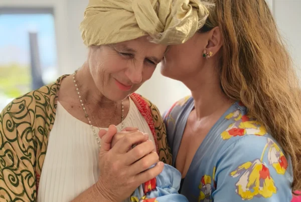 Two women holding hands; one whispers into the other’s ear while eyes close.
