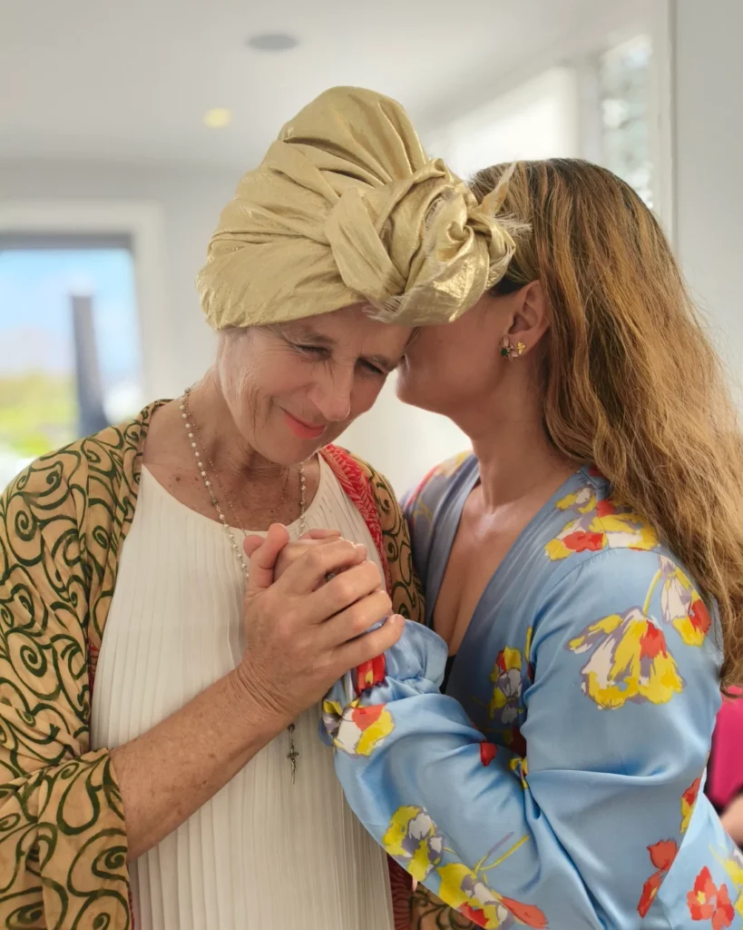 Two women holding hands; one whispers into the other’s ear while eyes close.